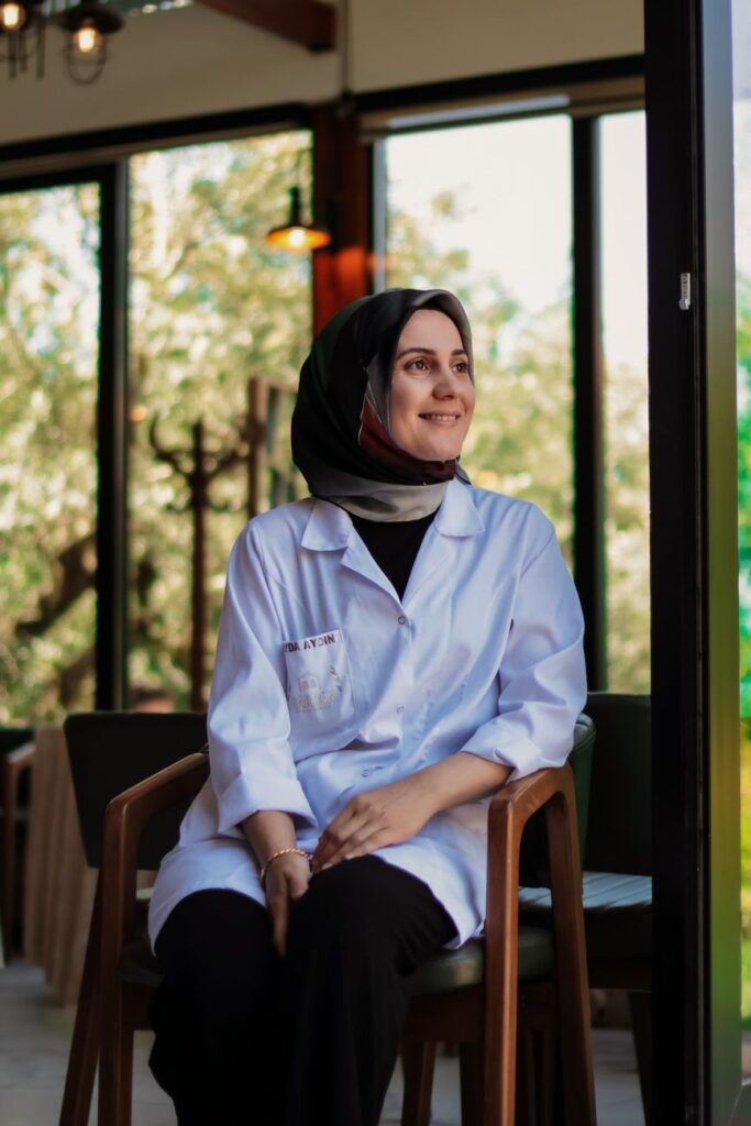 A smiling woman in a hijab and lab coat sits indoors in a sunlit setting.