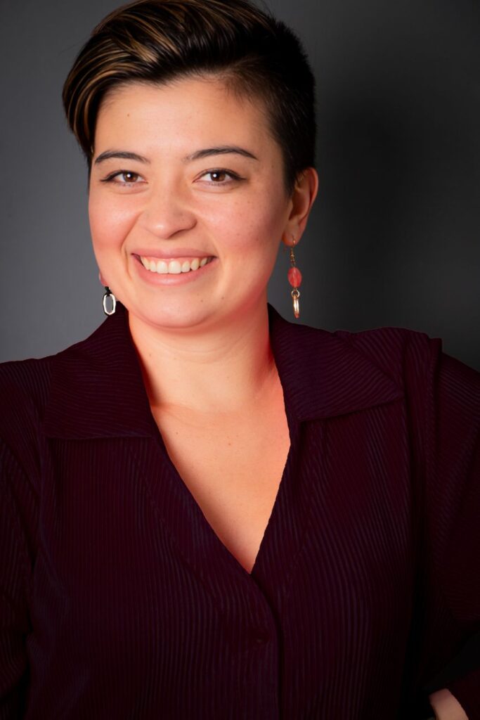 Close-up portrait of a smiling woman in formal attire, captured in a studio setting with a dark background.