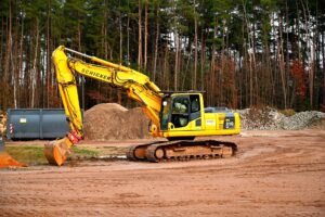 A yellow excavator at work in a forested construction site, surrounded by dirt and gravel.