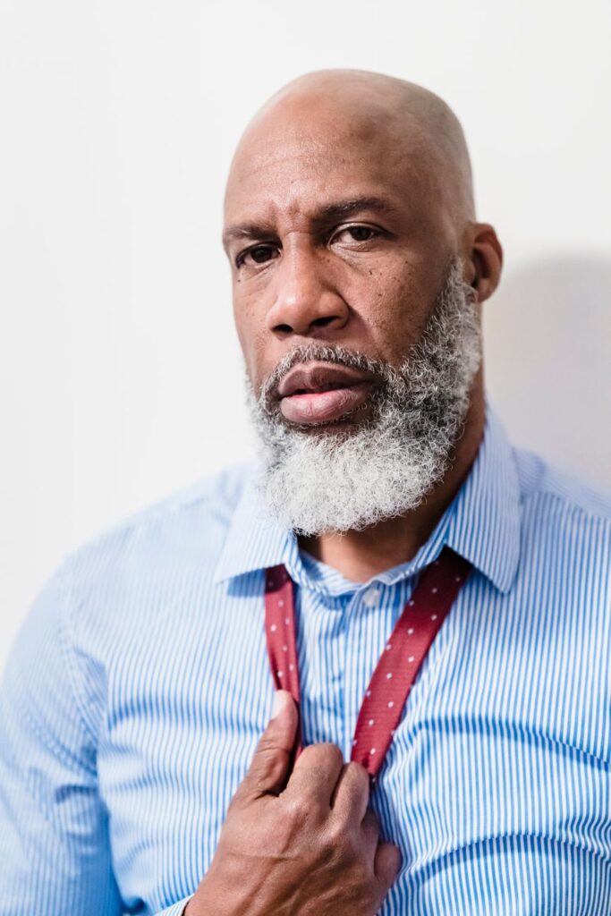 Portrait of a bald African American man with a gray beard adjusting his red tie against a white background.
